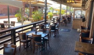 A vibrant dog park bathed in sunlight features abundant greenery with mature trees, offering ample shade for leisurely outdoor activities. In the distance is a well-equipped playground that presents an additional activity area. An inviting picnic table spreads out nearby, ideal for enjoying meals al fresco or taking relaxing breaks from play. A neatly-placed trash bin aids in maintaining the park's cleanliness for everyone's enjoyment. Dominating the scene is a generously wide dirt path gracefully curving through the expanse of grass, promising exploratory walks and lively fetch games.