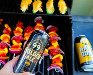 A vibrant dog park bathed in sunlight features abundant greenery with mature trees, offering ample shade for leisurely outdoor activities. In the distance is a well-equipped playground that presents an additional activity area. An inviting picnic table spreads out nearby, ideal for enjoying meals al fresco or taking relaxing breaks from play. A neatly-placed trash bin aids in maintaining the park's cleanliness for everyone's enjoyment. Dominating the scene is a generously wide dirt path gracefully curving through the expanse of grass, promising exploratory walks and lively fetch games.