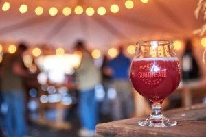 A vibrant dog park bathed in sunlight features abundant greenery with mature trees, offering ample shade for leisurely outdoor activities. In the distance is a well-equipped playground that presents an additional activity area. An inviting picnic table spreads out nearby, ideal for enjoying meals al fresco or taking relaxing breaks from play. A neatly-placed trash bin aids in maintaining the park's cleanliness for everyone's enjoyment. Dominating the scene is a generously wide dirt path gracefully curving through the expanse of grass, promising exploratory walks and lively fetch games.