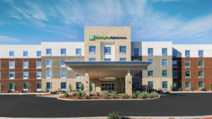 A vibrant dog park bathed in sunlight features abundant greenery with mature trees, offering ample shade for leisurely outdoor activities. In the distance is a well-equipped playground that presents an additional activity area. An inviting picnic table spreads out nearby, ideal for enjoying meals al fresco or taking relaxing breaks from play. A neatly-placed trash bin aids in maintaining the park's cleanliness for everyone's enjoyment. Dominating the scene is a generously wide dirt path gracefully curving through the expanse of grass, promising exploratory walks and lively fetch games.