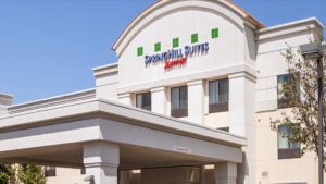 A vibrant dog park bathed in sunlight features abundant greenery with mature trees, offering ample shade for leisurely outdoor activities. In the distance is a well-equipped playground that presents an additional activity area. An inviting picnic table spreads out nearby, ideal for enjoying meals al fresco or taking relaxing breaks from play. A neatly-placed trash bin aids in maintaining the park's cleanliness for everyone's enjoyment. Dominating the scene is a generously wide dirt path gracefully curving through the expanse of grass, promising exploratory walks and lively fetch games.