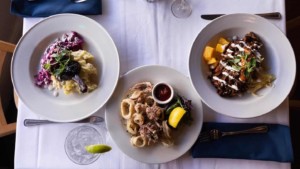 A vibrant dog park bathed in sunlight features abundant greenery with mature trees, offering ample shade for leisurely outdoor activities. In the distance is a well-equipped playground that presents an additional activity area. An inviting picnic table spreads out nearby, ideal for enjoying meals al fresco or taking relaxing breaks from play. A neatly-placed trash bin aids in maintaining the park's cleanliness for everyone's enjoyment. Dominating the scene is a generously wide dirt path gracefully curving through the expanse of grass, promising exploratory walks and lively fetch games.