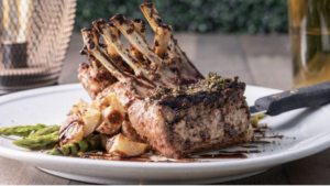 A vibrant dog park bathed in sunlight features abundant greenery with mature trees, offering ample shade for leisurely outdoor activities. In the distance is a well-equipped playground that presents an additional activity area. An inviting picnic table spreads out nearby, ideal for enjoying meals al fresco or taking relaxing breaks from play. A neatly-placed trash bin aids in maintaining the park's cleanliness for everyone's enjoyment. Dominating the scene is a generously wide dirt path gracefully curving through the expanse of grass, promising exploratory walks and lively fetch games.