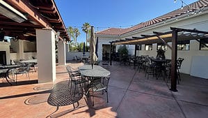 A vibrant dog park bathed in sunlight features abundant greenery with mature trees, offering ample shade for leisurely outdoor activities. In the distance is a well-equipped playground that presents an additional activity area. An inviting picnic table spreads out nearby, ideal for enjoying meals al fresco or taking relaxing breaks from play. A neatly-placed trash bin aids in maintaining the park's cleanliness for everyone's enjoyment. Dominating the scene is a generously wide dirt path gracefully curving through the expanse of grass, promising exploratory walks and lively fetch games.