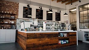 A vibrant dog park bathed in sunlight features abundant greenery with mature trees, offering ample shade for leisurely outdoor activities. In the distance is a well-equipped playground that presents an additional activity area. An inviting picnic table spreads out nearby, ideal for enjoying meals al fresco or taking relaxing breaks from play. A neatly-placed trash bin aids in maintaining the park's cleanliness for everyone's enjoyment. Dominating the scene is a generously wide dirt path gracefully curving through the expanse of grass, promising exploratory walks and lively fetch games.