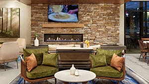 A vibrant dog park bathed in sunlight features abundant greenery with mature trees, offering ample shade for leisurely outdoor activities. In the distance is a well-equipped playground that presents an additional activity area. An inviting picnic table spreads out nearby, ideal for enjoying meals al fresco or taking relaxing breaks from play. A neatly-placed trash bin aids in maintaining the park's cleanliness for everyone's enjoyment. Dominating the scene is a generously wide dirt path gracefully curving through the expanse of grass, promising exploratory walks and lively fetch games.