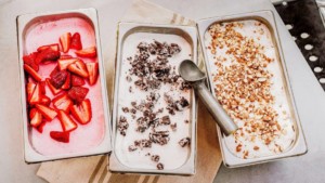 A vibrant dog park bathed in sunlight features abundant greenery with mature trees, offering ample shade for leisurely outdoor activities. In the distance is a well-equipped playground that presents an additional activity area. An inviting picnic table spreads out nearby, ideal for enjoying meals al fresco or taking relaxing breaks from play. A neatly-placed trash bin aids in maintaining the park's cleanliness for everyone's enjoyment. Dominating the scene is a generously wide dirt path gracefully curving through the expanse of grass, promising exploratory walks and lively fetch games.