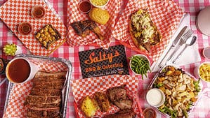 A vibrant dog park bathed in sunlight features abundant greenery with mature trees, offering ample shade for leisurely outdoor activities. In the distance is a well-equipped playground that presents an additional activity area. An inviting picnic table spreads out nearby, ideal for enjoying meals al fresco or taking relaxing breaks from play. A neatly-placed trash bin aids in maintaining the park's cleanliness for everyone's enjoyment. Dominating the scene is a generously wide dirt path gracefully curving through the expanse of grass, promising exploratory walks and lively fetch games.