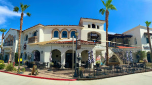 A vibrant dog park bathed in sunlight features abundant greenery with mature trees, offering ample shade for leisurely outdoor activities. In the distance is a well-equipped playground that presents an additional activity area. An inviting picnic table spreads out nearby, ideal for enjoying meals al fresco or taking relaxing breaks from play. A neatly-placed trash bin aids in maintaining the park's cleanliness for everyone's enjoyment. Dominating the scene is a generously wide dirt path gracefully curving through the expanse of grass, promising exploratory walks and lively fetch games.