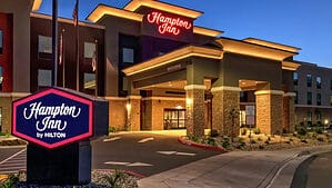 A vibrant dog park bathed in sunlight features abundant greenery with mature trees, offering ample shade for leisurely outdoor activities. In the distance is a well-equipped playground that presents an additional activity area. An inviting picnic table spreads out nearby, ideal for enjoying meals al fresco or taking relaxing breaks from play. A neatly-placed trash bin aids in maintaining the park's cleanliness for everyone's enjoyment. Dominating the scene is a generously wide dirt path gracefully curving through the expanse of grass, promising exploratory walks and lively fetch games.