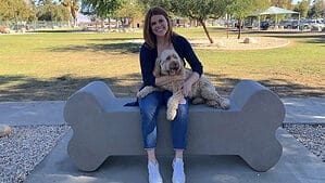 A vibrant dog park bathed in sunlight features abundant greenery with mature trees, offering ample shade for leisurely outdoor activities. In the distance is a well-equipped playground that presents an additional activity area. An inviting picnic table spreads out nearby, ideal for enjoying meals al fresco or taking relaxing breaks from play. A neatly-placed trash bin aids in maintaining the park's cleanliness for everyone's enjoyment. Dominating the scene is a generously wide dirt path gracefully curving through the expanse of grass, promising exploratory walks and lively fetch games.