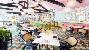 A vibrant dog park bathed in sunlight features abundant greenery with mature trees, offering ample shade for leisurely outdoor activities. In the distance is a well-equipped playground that presents an additional activity area. An inviting picnic table spreads out nearby, ideal for enjoying meals al fresco or taking relaxing breaks from play. A neatly-placed trash bin aids in maintaining the park's cleanliness for everyone's enjoyment. Dominating the scene is a generously wide dirt path gracefully curving through the expanse of grass, promising exploratory walks and lively fetch games.