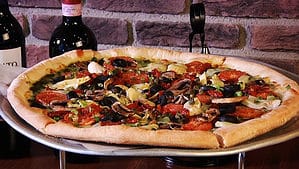 A vibrant dog park bathed in sunlight features abundant greenery with mature trees, offering ample shade for leisurely outdoor activities. In the distance is a well-equipped playground that presents an additional activity area. An inviting picnic table spreads out nearby, ideal for enjoying meals al fresco or taking relaxing breaks from play. A neatly-placed trash bin aids in maintaining the park's cleanliness for everyone's enjoyment. Dominating the scene is a generously wide dirt path gracefully curving through the expanse of grass, promising exploratory walks and lively fetch games.