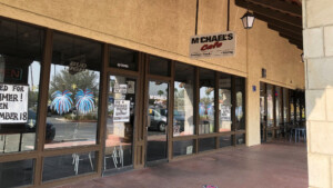 A vibrant dog park bathed in sunlight features abundant greenery with mature trees, offering ample shade for leisurely outdoor activities. In the distance is a well-equipped playground that presents an additional activity area. An inviting picnic table spreads out nearby, ideal for enjoying meals al fresco or taking relaxing breaks from play. A neatly-placed trash bin aids in maintaining the park's cleanliness for everyone's enjoyment. Dominating the scene is a generously wide dirt path gracefully curving through the expanse of grass, promising exploratory walks and lively fetch games.