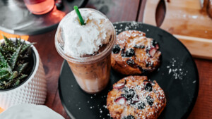 A vibrant dog park bathed in sunlight features abundant greenery with mature trees, offering ample shade for leisurely outdoor activities. In the distance is a well-equipped playground that presents an additional activity area. An inviting picnic table spreads out nearby, ideal for enjoying meals al fresco or taking relaxing breaks from play. A neatly-placed trash bin aids in maintaining the park's cleanliness for everyone's enjoyment. Dominating the scene is a generously wide dirt path gracefully curving through the expanse of grass, promising exploratory walks and lively fetch games.