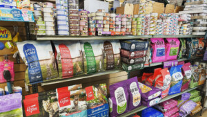 A vibrant dog park bathed in sunlight features abundant greenery with mature trees, offering ample shade for leisurely outdoor activities. In the distance is a well-equipped playground that presents an additional activity area. An inviting picnic table spreads out nearby, ideal for enjoying meals al fresco or taking relaxing breaks from play. A neatly-placed trash bin aids in maintaining the park's cleanliness for everyone's enjoyment. Dominating the scene is a generously wide dirt path gracefully curving through the expanse of grass, promising exploratory walks and lively fetch games.