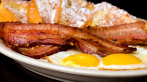 A vibrant dog park bathed in sunlight features abundant greenery with mature trees, offering ample shade for leisurely outdoor activities. In the distance is a well-equipped playground that presents an additional activity area. An inviting picnic table spreads out nearby, ideal for enjoying meals al fresco or taking relaxing breaks from play. A neatly-placed trash bin aids in maintaining the park's cleanliness for everyone's enjoyment. Dominating the scene is a generously wide dirt path gracefully curving through the expanse of grass, promising exploratory walks and lively fetch games.