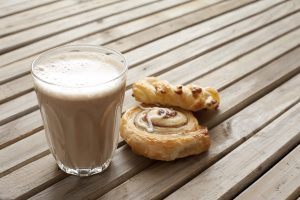 A vibrant dog park bathed in sunlight features abundant greenery with mature trees, offering ample shade for leisurely outdoor activities. In the distance is a well-equipped playground that presents an additional activity area. An inviting picnic table spreads out nearby, ideal for enjoying meals al fresco or taking relaxing breaks from play. A neatly-placed trash bin aids in maintaining the park's cleanliness for everyone's enjoyment. Dominating the scene is a generously wide dirt path gracefully curving through the expanse of grass, promising exploratory walks and lively fetch games.