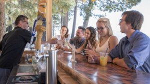 A vibrant dog park bathed in sunlight features abundant greenery with mature trees, offering ample shade for leisurely outdoor activities. In the distance is a well-equipped playground that presents an additional activity area. An inviting picnic table spreads out nearby, ideal for enjoying meals al fresco or taking relaxing breaks from play. A neatly-placed trash bin aids in maintaining the park's cleanliness for everyone's enjoyment. Dominating the scene is a generously wide dirt path gracefully curving through the expanse of grass, promising exploratory walks and lively fetch games.