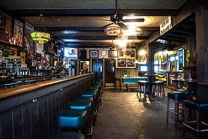 A vibrant dog park bathed in sunlight features abundant greenery with mature trees, offering ample shade for leisurely outdoor activities. In the distance is a well-equipped playground that presents an additional activity area. An inviting picnic table spreads out nearby, ideal for enjoying meals al fresco or taking relaxing breaks from play. A neatly-placed trash bin aids in maintaining the park's cleanliness for everyone's enjoyment. Dominating the scene is a generously wide dirt path gracefully curving through the expanse of grass, promising exploratory walks and lively fetch games.
