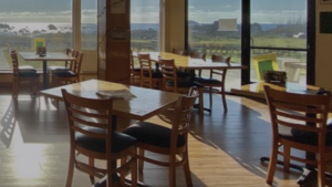 A vibrant dog park bathed in sunlight features abundant greenery with mature trees, offering ample shade for leisurely outdoor activities. In the distance is a well-equipped playground that presents an additional activity area. An inviting picnic table spreads out nearby, ideal for enjoying meals al fresco or taking relaxing breaks from play. A neatly-placed trash bin aids in maintaining the park's cleanliness for everyone's enjoyment. Dominating the scene is a generously wide dirt path gracefully curving through the expanse of grass, promising exploratory walks and lively fetch games.