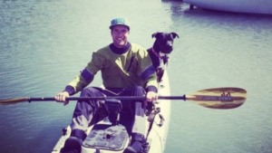 A vibrant dog park bathed in sunlight features abundant greenery with mature trees, offering ample shade for leisurely outdoor activities. In the distance is a well-equipped playground that presents an additional activity area. An inviting picnic table spreads out nearby, ideal for enjoying meals al fresco or taking relaxing breaks from play. A neatly-placed trash bin aids in maintaining the park's cleanliness for everyone's enjoyment. Dominating the scene is a generously wide dirt path gracefully curving through the expanse of grass, promising exploratory walks and lively fetch games.