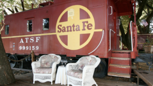 A vibrant dog park bathed in sunlight features abundant greenery with mature trees, offering ample shade for leisurely outdoor activities. In the distance is a well-equipped playground that presents an additional activity area. An inviting picnic table spreads out nearby, ideal for enjoying meals al fresco or taking relaxing breaks from play. A neatly-placed trash bin aids in maintaining the park's cleanliness for everyone's enjoyment. Dominating the scene is a generously wide dirt path gracefully curving through the expanse of grass, promising exploratory walks and lively fetch games.