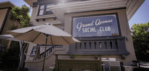 A vibrant dog park bathed in sunlight features abundant greenery with mature trees, offering ample shade for leisurely outdoor activities. In the distance is a well-equipped playground that presents an additional activity area. An inviting picnic table spreads out nearby, ideal for enjoying meals al fresco or taking relaxing breaks from play. A neatly-placed trash bin aids in maintaining the park's cleanliness for everyone's enjoyment. Dominating the scene is a generously wide dirt path gracefully curving through the expanse of grass, promising exploratory walks and lively fetch games.