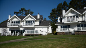 A vibrant dog park bathed in sunlight features abundant greenery with mature trees, offering ample shade for leisurely outdoor activities. In the distance is a well-equipped playground that presents an additional activity area. An inviting picnic table spreads out nearby, ideal for enjoying meals al fresco or taking relaxing breaks from play. A neatly-placed trash bin aids in maintaining the park's cleanliness for everyone's enjoyment. Dominating the scene is a generously wide dirt path gracefully curving through the expanse of grass, promising exploratory walks and lively fetch games.