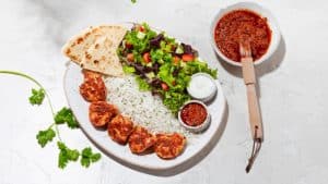 A vibrant dog park bathed in sunlight features abundant greenery with mature trees, offering ample shade for leisurely outdoor activities. In the distance is a well-equipped playground that presents an additional activity area. An inviting picnic table spreads out nearby, ideal for enjoying meals al fresco or taking relaxing breaks from play. A neatly-placed trash bin aids in maintaining the park's cleanliness for everyone's enjoyment. Dominating the scene is a generously wide dirt path gracefully curving through the expanse of grass, promising exploratory walks and lively fetch games.