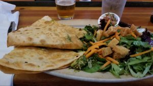 A vibrant dog park bathed in sunlight features abundant greenery with mature trees, offering ample shade for leisurely outdoor activities. In the distance is a well-equipped playground that presents an additional activity area. An inviting picnic table spreads out nearby, ideal for enjoying meals al fresco or taking relaxing breaks from play. A neatly-placed trash bin aids in maintaining the park's cleanliness for everyone's enjoyment. Dominating the scene is a generously wide dirt path gracefully curving through the expanse of grass, promising exploratory walks and lively fetch games.