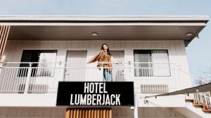 A vibrant dog park bathed in sunlight features abundant greenery with mature trees, offering ample shade for leisurely outdoor activities. In the distance is a well-equipped playground that presents an additional activity area. An inviting picnic table spreads out nearby, ideal for enjoying meals al fresco or taking relaxing breaks from play. A neatly-placed trash bin aids in maintaining the park's cleanliness for everyone's enjoyment. Dominating the scene is a generously wide dirt path gracefully curving through the expanse of grass, promising exploratory walks and lively fetch games.