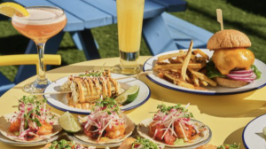 A vibrant dog park bathed in sunlight features abundant greenery with mature trees, offering ample shade for leisurely outdoor activities. In the distance is a well-equipped playground that presents an additional activity area. An inviting picnic table spreads out nearby, ideal for enjoying meals al fresco or taking relaxing breaks from play. A neatly-placed trash bin aids in maintaining the park's cleanliness for everyone's enjoyment. Dominating the scene is a generously wide dirt path gracefully curving through the expanse of grass, promising exploratory walks and lively fetch games.