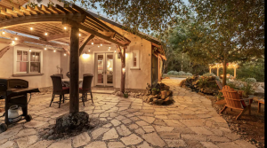 A vibrant dog park bathed in sunlight features abundant greenery with mature trees, offering ample shade for leisurely outdoor activities. In the distance is a well-equipped playground that presents an additional activity area. An inviting picnic table spreads out nearby, ideal for enjoying meals al fresco or taking relaxing breaks from play. A neatly-placed trash bin aids in maintaining the park's cleanliness for everyone's enjoyment. Dominating the scene is a generously wide dirt path gracefully curving through the expanse of grass, promising exploratory walks and lively fetch games.