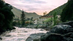 A vibrant dog park bathed in sunlight features abundant greenery with mature trees, offering ample shade for leisurely outdoor activities. In the distance is a well-equipped playground that presents an additional activity area. An inviting picnic table spreads out nearby, ideal for enjoying meals al fresco or taking relaxing breaks from play. A neatly-placed trash bin aids in maintaining the park's cleanliness for everyone's enjoyment. Dominating the scene is a generously wide dirt path gracefully curving through the expanse of grass, promising exploratory walks and lively fetch games.