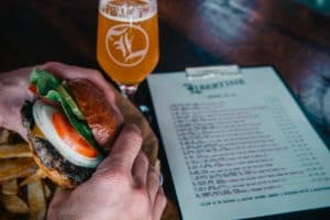 A vibrant dog park bathed in sunlight features abundant greenery with mature trees, offering ample shade for leisurely outdoor activities. In the distance is a well-equipped playground that presents an additional activity area. An inviting picnic table spreads out nearby, ideal for enjoying meals al fresco or taking relaxing breaks from play. A neatly-placed trash bin aids in maintaining the park's cleanliness for everyone's enjoyment. Dominating the scene is a generously wide dirt path gracefully curving through the expanse of grass, promising exploratory walks and lively fetch games.