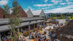 A vibrant dog park bathed in sunlight features abundant greenery with mature trees, offering ample shade for leisurely outdoor activities. In the distance is a well-equipped playground that presents an additional activity area. An inviting picnic table spreads out nearby, ideal for enjoying meals al fresco or taking relaxing breaks from play. A neatly-placed trash bin aids in maintaining the park's cleanliness for everyone's enjoyment. Dominating the scene is a generously wide dirt path gracefully curving through the expanse of grass, promising exploratory walks and lively fetch games.