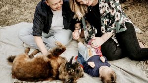A vibrant dog park bathed in sunlight features abundant greenery with mature trees, offering ample shade for leisurely outdoor activities. In the distance is a well-equipped playground that presents an additional activity area. An inviting picnic table spreads out nearby, ideal for enjoying meals al fresco or taking relaxing breaks from play. A neatly-placed trash bin aids in maintaining the park's cleanliness for everyone's enjoyment. Dominating the scene is a generously wide dirt path gracefully curving through the expanse of grass, promising exploratory walks and lively fetch games.