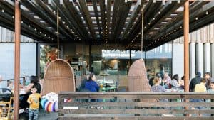 A vibrant dog park bathed in sunlight features abundant greenery with mature trees, offering ample shade for leisurely outdoor activities. In the distance is a well-equipped playground that presents an additional activity area. An inviting picnic table spreads out nearby, ideal for enjoying meals al fresco or taking relaxing breaks from play. A neatly-placed trash bin aids in maintaining the park's cleanliness for everyone's enjoyment. Dominating the scene is a generously wide dirt path gracefully curving through the expanse of grass, promising exploratory walks and lively fetch games.