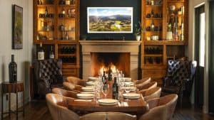 A vibrant dog park bathed in sunlight features abundant greenery with mature trees, offering ample shade for leisurely outdoor activities. In the distance is a well-equipped playground that presents an additional activity area. An inviting picnic table spreads out nearby, ideal for enjoying meals al fresco or taking relaxing breaks from play. A neatly-placed trash bin aids in maintaining the park's cleanliness for everyone's enjoyment. Dominating the scene is a generously wide dirt path gracefully curving through the expanse of grass, promising exploratory walks and lively fetch games.