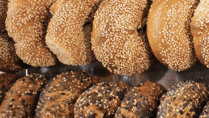 A vibrant dog park bathed in sunlight features abundant greenery with mature trees, offering ample shade for leisurely outdoor activities. In the distance is a well-equipped playground that presents an additional activity area. An inviting picnic table spreads out nearby, ideal for enjoying meals al fresco or taking relaxing breaks from play. A neatly-placed trash bin aids in maintaining the park's cleanliness for everyone's enjoyment. Dominating the scene is a generously wide dirt path gracefully curving through the expanse of grass, promising exploratory walks and lively fetch games.