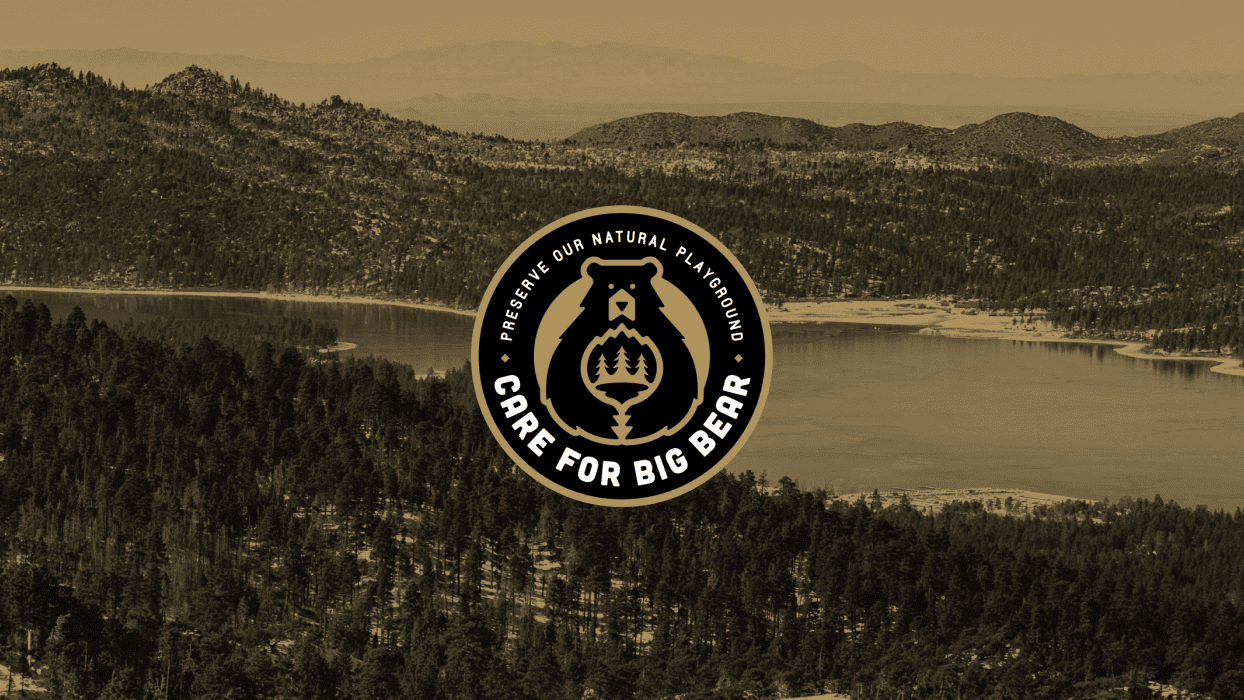 A vibrant dog park bathed in sunlight features abundant greenery with mature trees, offering ample shade for leisurely outdoor activities. In the distance is a well-equipped playground that presents an additional activity area. An inviting picnic table spreads out nearby, ideal for enjoying meals al fresco or taking relaxing breaks from play. A neatly-placed trash bin aids in maintaining the park's cleanliness for everyone's enjoyment. Dominating the scene is a generously wide dirt path gracefully curving through the expanse of grass, promising exploratory walks and lively fetch games.