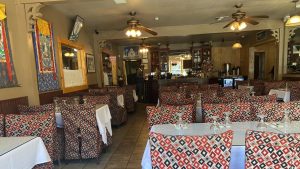 A vibrant dog park bathed in sunlight features abundant greenery with mature trees, offering ample shade for leisurely outdoor activities. In the distance is a well-equipped playground that presents an additional activity area. An inviting picnic table spreads out nearby, ideal for enjoying meals al fresco or taking relaxing breaks from play. A neatly-placed trash bin aids in maintaining the park's cleanliness for everyone's enjoyment. Dominating the scene is a generously wide dirt path gracefully curving through the expanse of grass, promising exploratory walks and lively fetch games.