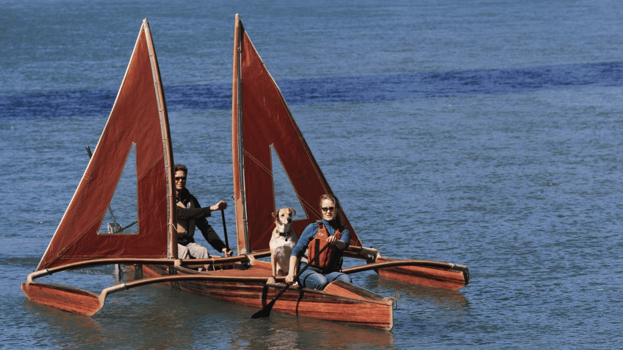 Two individuals accompanied by their dog, embark on a journey atop catamarans adorned with vibrant red sails.