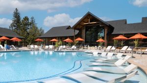 A vibrant dog park bathed in sunlight features abundant greenery with mature trees, offering ample shade for leisurely outdoor activities. In the distance is a well-equipped playground that presents an additional activity area. An inviting picnic table spreads out nearby, ideal for enjoying meals al fresco or taking relaxing breaks from play. A neatly-placed trash bin aids in maintaining the park's cleanliness for everyone's enjoyment. Dominating the scene is a generously wide dirt path gracefully curving through the expanse of grass, promising exploratory walks and lively fetch games.