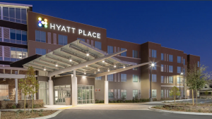 A vibrant dog park bathed in sunlight features abundant greenery with mature trees, offering ample shade for leisurely outdoor activities. In the distance is a well-equipped playground that presents an additional activity area. An inviting picnic table spreads out nearby, ideal for enjoying meals al fresco or taking relaxing breaks from play. A neatly-placed trash bin aids in maintaining the park's cleanliness for everyone's enjoyment. Dominating the scene is a generously wide dirt path gracefully curving through the expanse of grass, promising exploratory walks and lively fetch games.