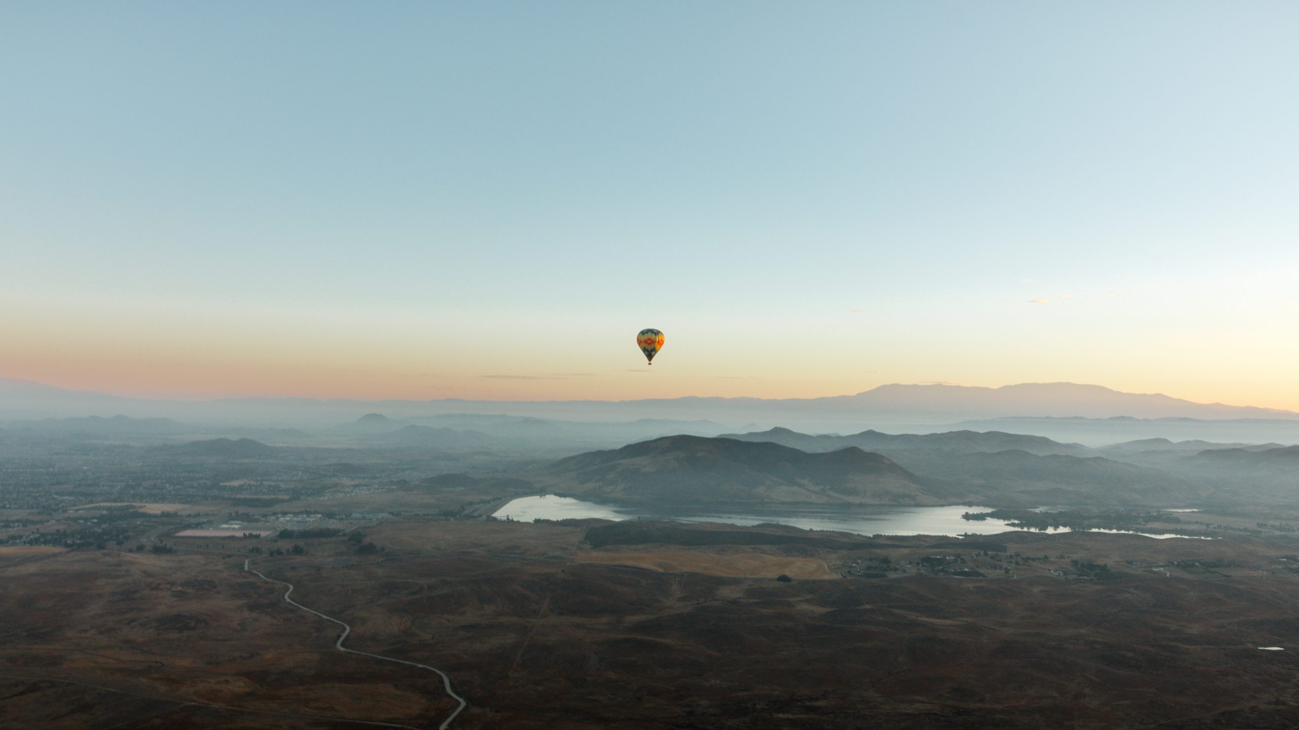 A vibrant dog park bathed in sunlight features abundant greenery with mature trees, offering ample shade for leisurely outdoor activities. In the distance is a well-equipped playground that presents an additional activity area. An inviting picnic table spreads out nearby, ideal for enjoying meals al fresco or taking relaxing breaks from play. A neatly-placed trash bin aids in maintaining the park's cleanliness for everyone's enjoyment. Dominating the scene is a generously wide dirt path gracefully curving through the expanse of grass, promising exploratory walks and lively fetch games.