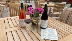 A vibrant dog park bathed in sunlight features abundant greenery with mature trees, offering ample shade for leisurely outdoor activities. In the distance is a well-equipped playground that presents an additional activity area. An inviting picnic table spreads out nearby, ideal for enjoying meals al fresco or taking relaxing breaks from play. A neatly-placed trash bin aids in maintaining the park's cleanliness for everyone's enjoyment. Dominating the scene is a generously wide dirt path gracefully curving through the expanse of grass, promising exploratory walks and lively fetch games.