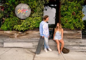 A vibrant dog park bathed in sunlight features abundant greenery with mature trees, offering ample shade for leisurely outdoor activities. In the distance is a well-equipped playground that presents an additional activity area. An inviting picnic table spreads out nearby, ideal for enjoying meals al fresco or taking relaxing breaks from play. A neatly-placed trash bin aids in maintaining the park's cleanliness for everyone's enjoyment. Dominating the scene is a generously wide dirt path gracefully curving through the expanse of grass, promising exploratory walks and lively fetch games.