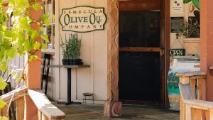 A vibrant dog park bathed in sunlight features abundant greenery with mature trees, offering ample shade for leisurely outdoor activities. In the distance is a well-equipped playground that presents an additional activity area. An inviting picnic table spreads out nearby, ideal for enjoying meals al fresco or taking relaxing breaks from play. A neatly-placed trash bin aids in maintaining the park's cleanliness for everyone's enjoyment. Dominating the scene is a generously wide dirt path gracefully curving through the expanse of grass, promising exploratory walks and lively fetch games.